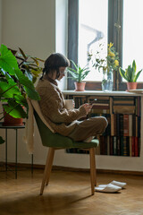 Lonely teen girl sitting with smartphone holding cup of tea near window surfing internet social...