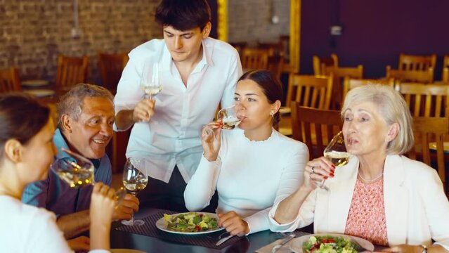 Cheerful elderly couple spending time with young friends in cozy restaurant. Man and woman having fun while talking and enjoying light dinner with wine at table