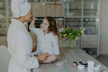 Curly girl touches mom's nose, in white robe, on cosmetic-filled table, preparing to do makeup. Cozy home interior.