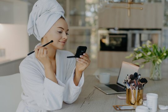 Beautiful woman applies makeup with brush, poses at home, in front of mirror. Desk with laptop, cosmetology tools, bathrobe. Self-care concept.