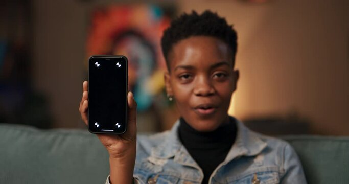 Tech enthusiast beta testerAfrican woman sitting in living room, holds her cellphone with a dark screen, gesturing with excitement, eagerly awaiting a new software update app release.