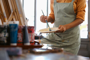Cropped image of female artist standing in front of an easel and dipping brush into color palette