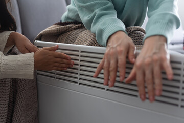 Mother and child warming hands near electric heater at home, closeup.