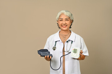 Senior woman doctor in white coat holding digital blood pressure monitor against brown background - Powered by Adobe