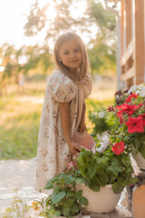  little girl with blonde hair is watering flowers on the veranda in summer at sunset