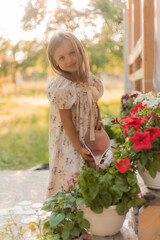  little girl with blonde hair is watering flowers on the veranda in summer at sunset