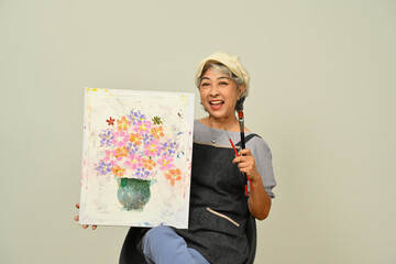 Happy senior woman 60s wearing apron holding canvas and brush over gray background