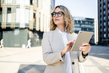 A blonde woman with glasses, a businesswoman in a jacket, goes to the fois and uses a tablet.