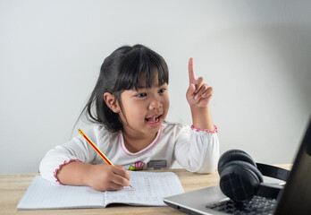 4-5 years old Asian girl wearing white shirt is studying online via computer using bluetooth headphones at home, distance education concept