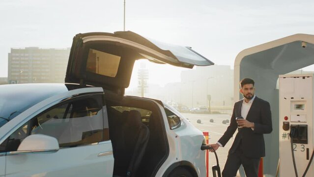 Indian man at station charging electric car. Arabian businessman in suit inserting power cord into white electric car while standing outdoors with takeaway coffee in one hand. Zero emission vehicle
