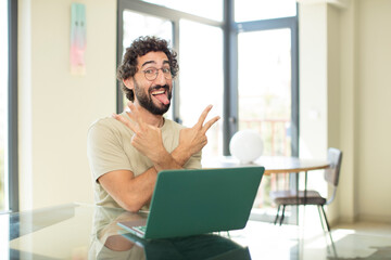 young adult bearded man with a laptop smiling and looking happy, friendly and satisfied, gesturing victory or peace with both hands