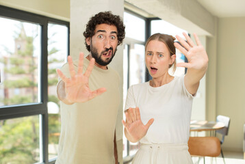 young adult couple feeling stupefied and scared, fearing something frightening, with hands open up...