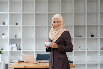 Asian Muslim business woman holding a coffee mug standing in the modern office. business people, diversity and office concept