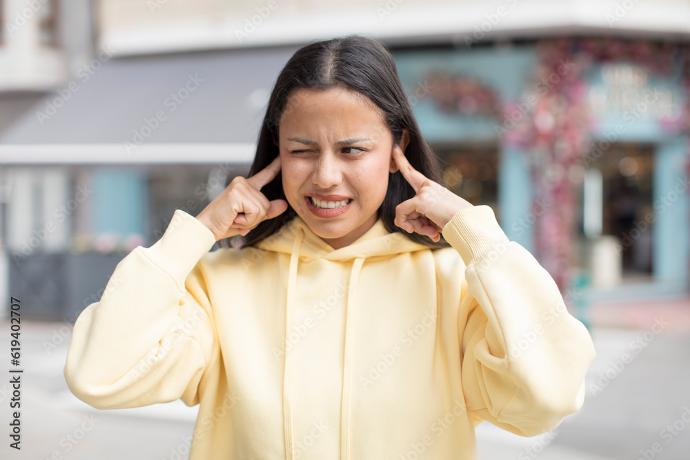 Wall mural pretty hispanic woman looking angry, stressed and annoyed, covering both ears to a deafening noise, sound or loud music