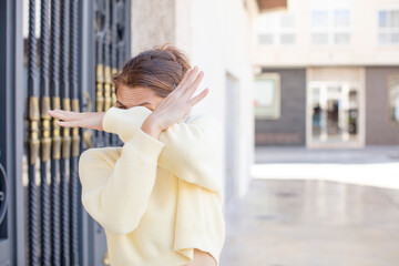 pretty young woman covering face with hand and putting other hand up front to stop camera, refusing...