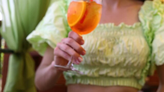 young woman, blonde girl, in restaurant outdoor terrace drinking lemonade or aperol spritz,with straw.glass on table or next to curtains against summer sky.refreshing beverage slice of lemon or orange