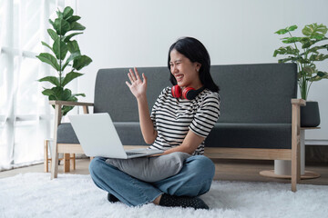 Portrait of happy attractive asian woman and having video call, waving hand at laptop, having online meeting or conversation while relaxing at home