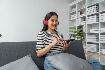 Attractive happy young Asian woman listening music with headphones from music application on laptop and telephone, sitting on sofa