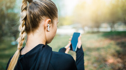 Young smiling girl making sport and running in the park using her phone to listen the music with wireless headphones on sunset in the city watching the screen