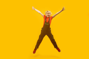 Fototapeta na wymiar Portrait of schoolboy in overalls bouncing on yellow background. Blond boy jumps high with his hands up. Copy space