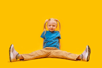 Caucasian little fair-haired sport girl with two ponytails wears blue T-shirt and yellow pants sits...