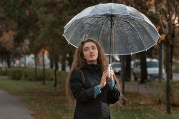 Beautiful girl with brown hair walks in autumn park. Young woman with transparent umbrella to see is raining