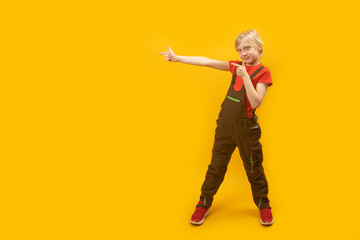 Blond boy wears jumpsuit and red T-shirt stands with his legs spread wide and pointing his index fingers on side. Copy space.