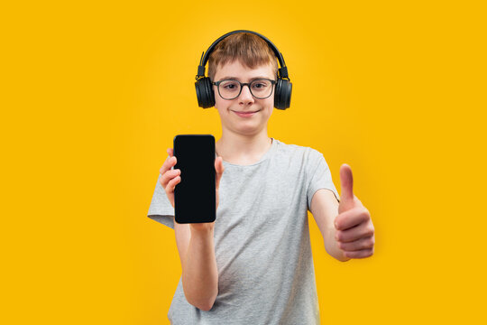 School boy wearing glasses and headphones holds smartphone with blank screen and shows thumbs up. Apps for listening audiobook or music. Copy space, mock up.