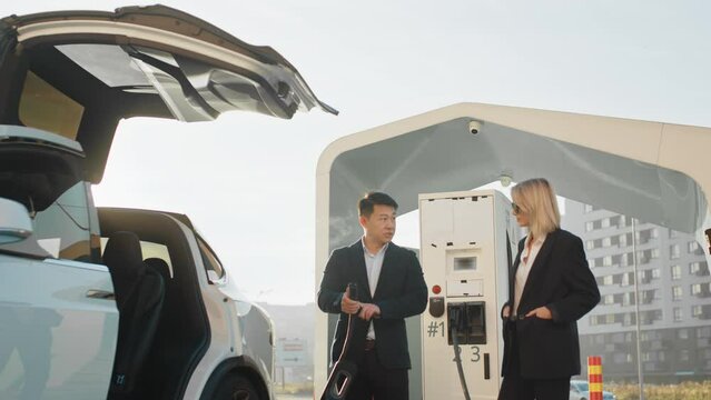 Transport and purchase concept. Stylish caucasian woman talking with asian car dealer while buying new electric vehicle. Multiracial people standing near moden auto on charging station. Electric car.