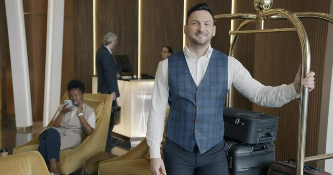 Young man porter is standing in hotel lobby with baggage cart smiling and looking at camera while people guests and staff are moving in background.