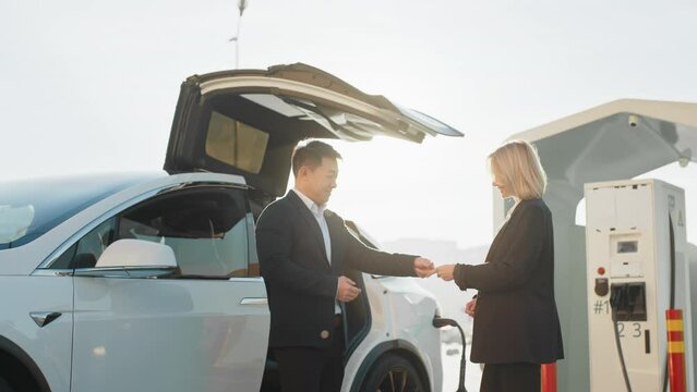 The seller hands over the keys to the new car. Female customer of dealership shaking hands with competent consultant while buying new electric auto. Woman and asian man standing together near car