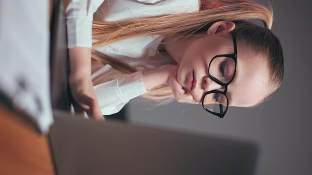 Vertical video of dreamy preteen girl dressed in white blouse and black glasses sitting still in office chair and slowly typing on keyboard. Blue-eyed child holding hand under chin and thinking.