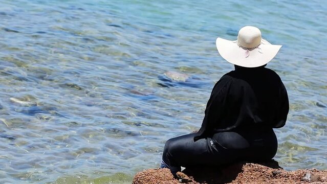 Muslim woman back view wearing black muslim dress and a hat (unrecognizable) sitting by the sea in Egypt