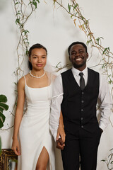 Vertical portrait of elegant black couple as bride and groom standing against wall decorated with...