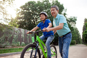 Obraz premium asian dad teaches son to ride bike, korean senior helps child, boy in helmet is actively relaxing with his father