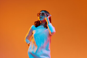 Portrait of young beautiful girl in casual clothes and trendy sunglasses posing against orange studio background in neon light. Concept of human emotions, youth, feelings, fashion, lifestyle, ad