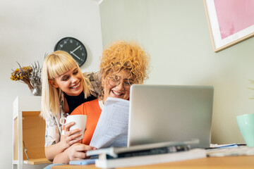 Lesbian couple laughing and hugging while using laptop together at home.