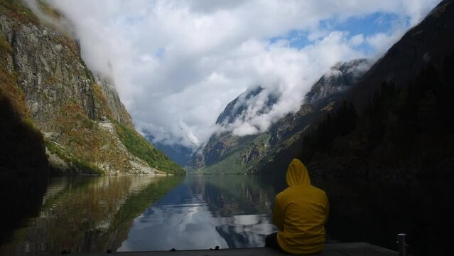 Young man in yellow rain coat enjoying the view of Norways Fjord landscape. impressive mountain reflecting in a calm lake