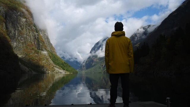 Young man in yellow rain coat enjoying the view of Norways Fjord landscape. impressive mountain reflecting in a calm lake