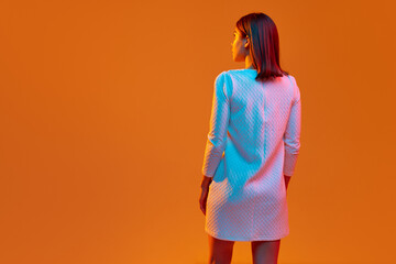 Back view image of young girl standing in white comfortable dress against orange studio background in neon light. Concept of human emotions, youth, feelings, fashion, lifestyle, ad