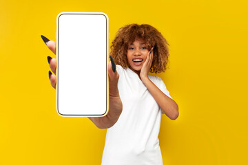 amazed curly american girl with braces in white t-shirt shows blank smartphone screen on yellow background