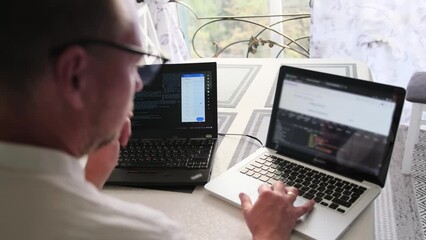 Focused IT specialist in glasses writes program code on two laptops while working at home near the window overlooking street, rear view. Programmer work in home office. Remote work arrangements.