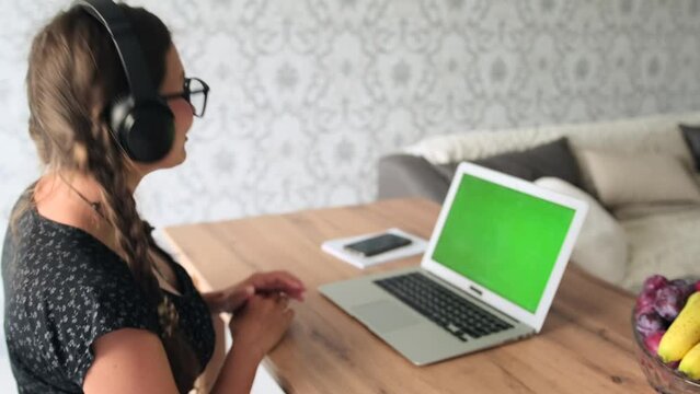 Positive beautiful caucasian woman in glasses and headphones with smile communicates via video call center operator talks to customer on online conference, work at home. Green screen.