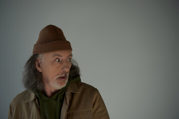 shocked hipster style man in beanie hat and brown jacket standing with open mouth and looking away on grey background, aging population lifestyle concept, copy space