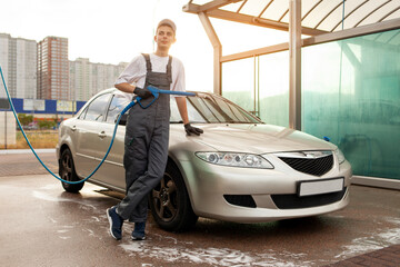 Fototapeta na wymiar young guy in uniform car wash worker stands in the background of car and holds hose