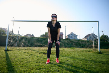 Attractive girl goalkeeper in black sports tight clothes standing near the goal, warming up, stretching in the morning at sunrise.