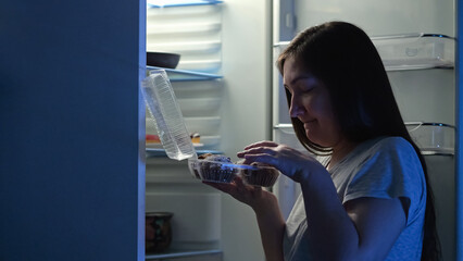 Hungry woman enjoys eating creamy doughnuts from fridge at nighttime. Long-haired lady grabs desserts from refrigerator to tasty at night