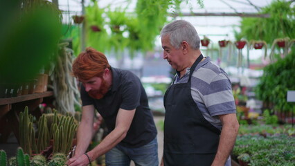 Older business store owner helping young client to buy plant at local Flower Shop. Senior man wearing apron assisting man with product purchase