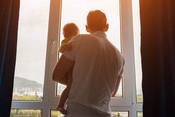 Father holding toddler daughter in arms opens curtains of panoramic window together. Parent and...