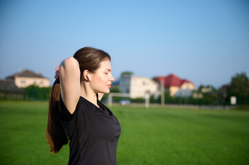 Portrait of young attractive fit girl in black sports tight clothes standing on green field before workout at sunrise. Caucasian female jogger runner on jogging track. Sport and health.